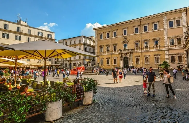 Cafe umbrellas and cobblestones in a sunlit Italian piazza, with people walking past a palazzo