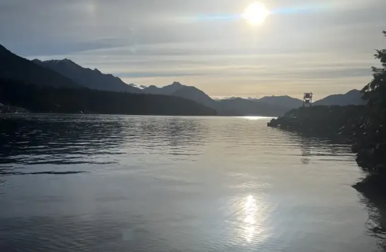 Calm Alaska Sitka bay reflecting sun with layered mountain silhouettes and overcast sky at dusk.