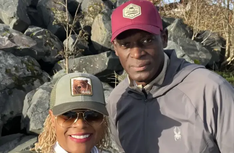 Two travelers wearing Alaska-branded caps pose near rocky shoreline in Sitka; Alaska on a sunny autumn day.