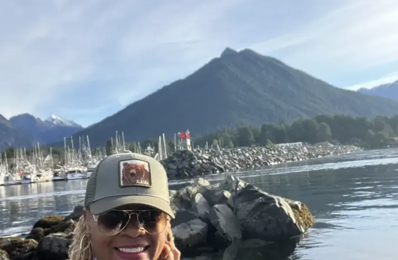 Smiling traveler in Alaska cap poses near Sitka harbor with a dramatic mountain peak and marina in the background.