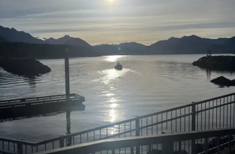 A boat glides through calm Alaska Sitka harbor waters at sunset; with mountain silhouettes and a glowing sun reflected on