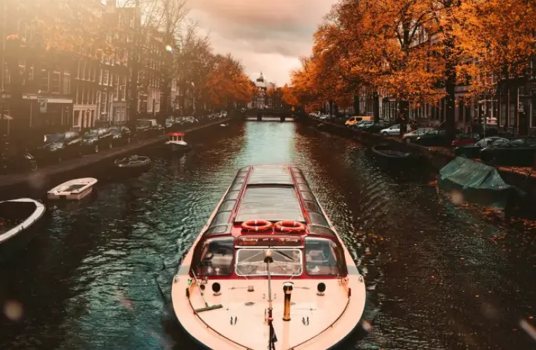 River cruise boat gliding through Amsterdam canal lined with golden autumn trees and historic canal houses at sunset.