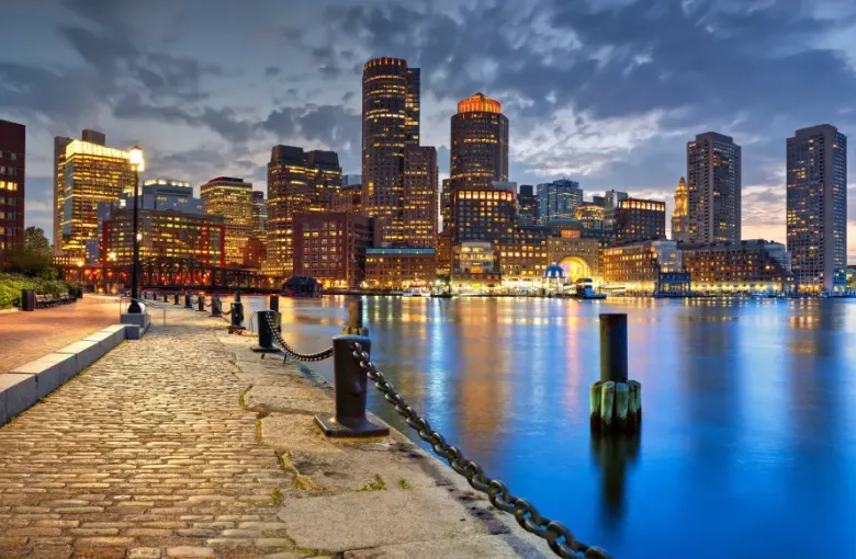 Boston Massachusetts waterfront skyline at dusk with illuminated skyscrapers reflecting on calm harbor water from a cobblestone promenade
