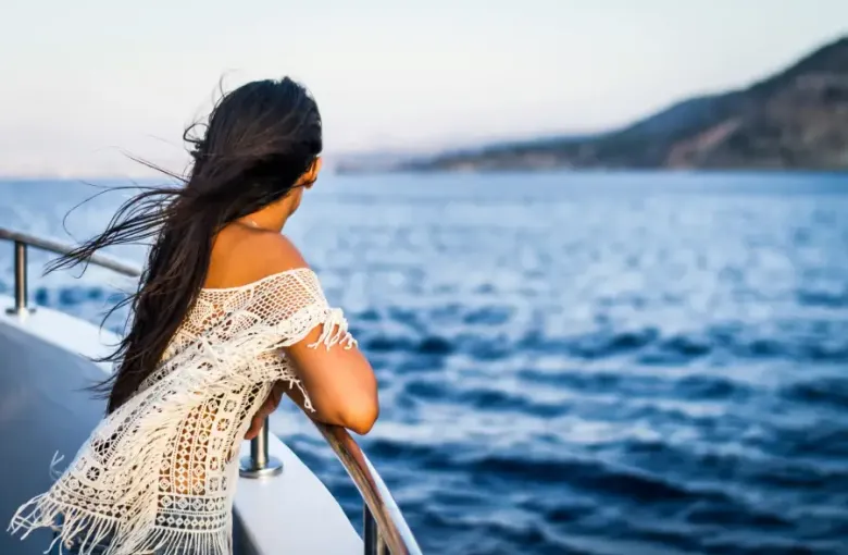 Woman in white lace top leaning on cruise ship railing; gazing at blue ocean waters with coastal hills at dusk.