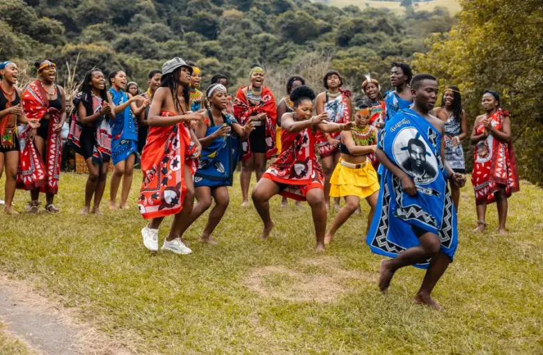 Group of young adults in vibrant traditional African attire performing a cultural dance outdoors on green grass; cruise-la...