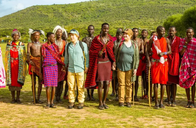 Two Western tourists stand among traditionally dressed Maasai community members during a cruise-land excursion in Kenya's...