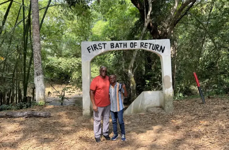 Two visitors pose under the 'First Bath of Return' arch at a heritage site during eat-mea-2024; surrounded by lush forest.
