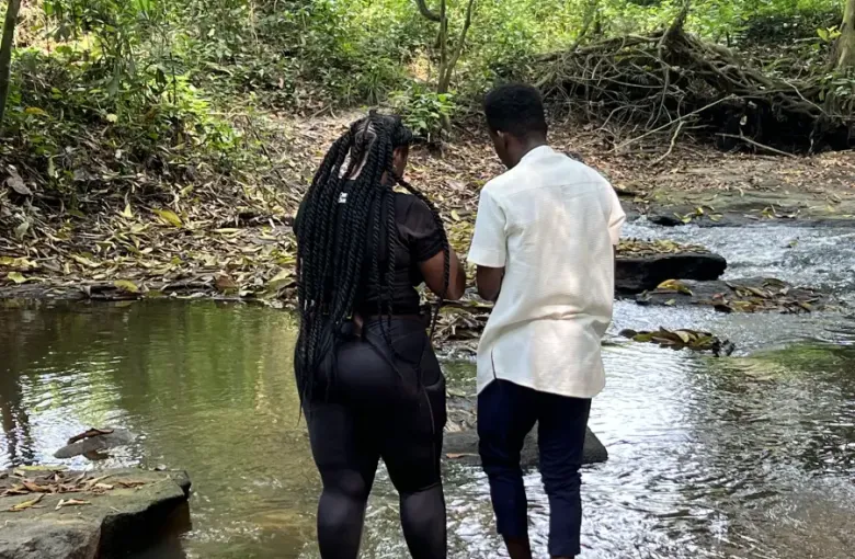 Two people stand barefoot at a forest stream during eat-mea-2024; surrounded by lush tropical greenery and rocks.