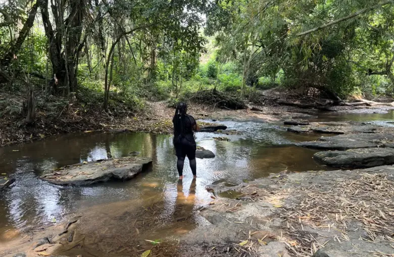 Woman in black wading barefoot through a shallow jungle stream on rocky terrain during EAT MEA 2024 nature excursion.