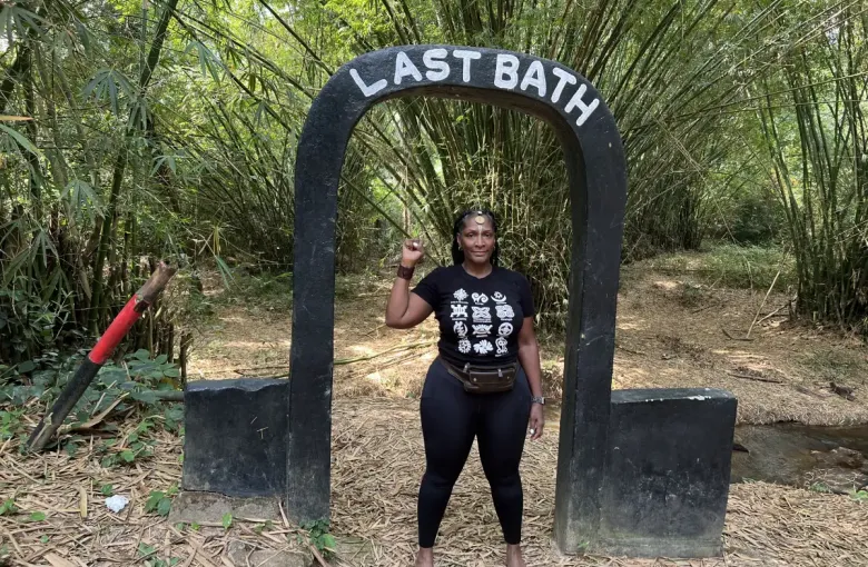 Woman raises fist under 'Last Bath' arch monument at eat-mea-2024; surrounded by bamboo forest in Ghana.