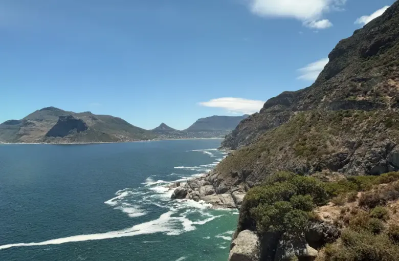 Dramatic rocky cliffs overlooking Hout Bay's turquoise waters with mountain backdrop; captured for eat-mea-2026 travel sho...