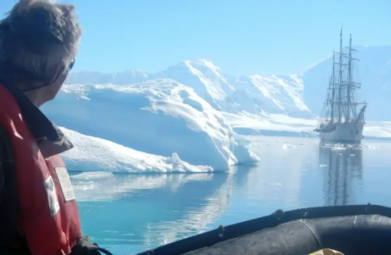 Traveler in red life vest on a Zodiac views a tall ship anchored among Antarctic icebergs during an expedition cruise.