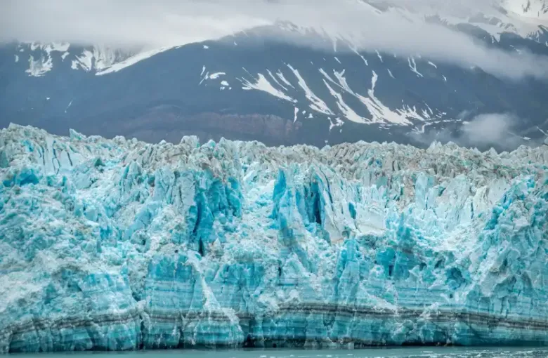 Tour boat with passengers navigating icy waters beside a massive blue glacier on an Alaska expedition cruise; mountains be