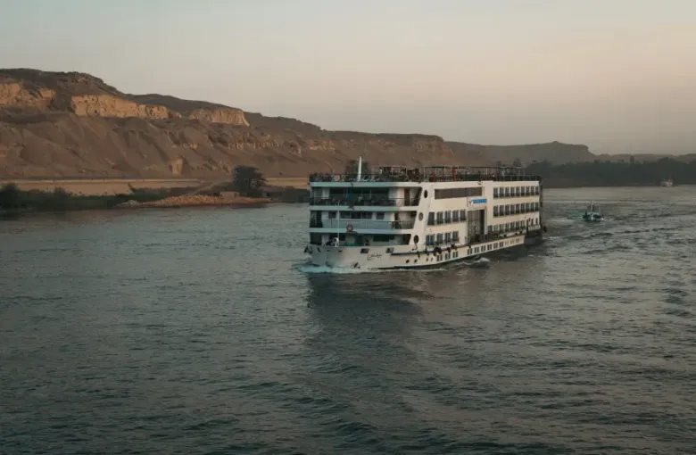 Multi-deck river cruise ship sailing the Nile at dusk with desert cliffs and palm trees in the background.