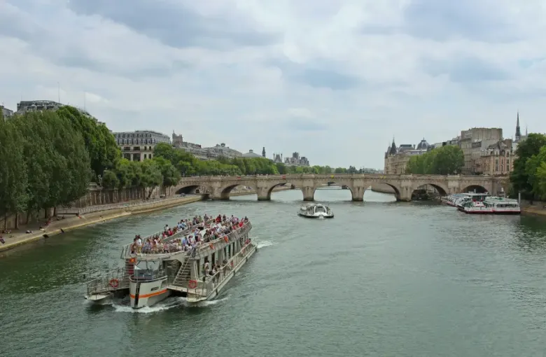 Packed river cruise boat on the Seine in Paris; with Pont Neuf arches and Haussmann buildings in the background.