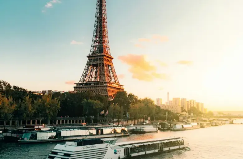 Paris Seine River cruise boat passing the Eiffel Tower at golden sunset; with city skyline in background.