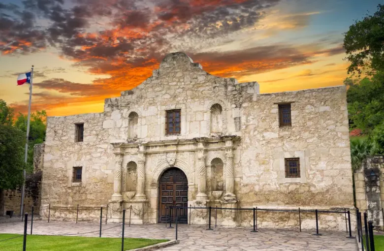 The Alamo's limestone facade at sunset in San Antonio Texas with the Texas flag flying and a dramatic orange sky