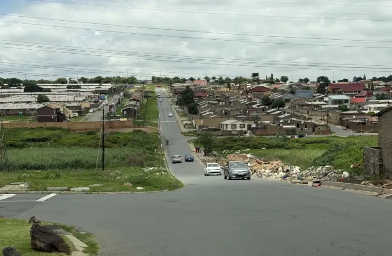 A long residential street in Johannesburg South Africa lined with modest brick homes; parked cars; and lush green verges under a cloudy sky.