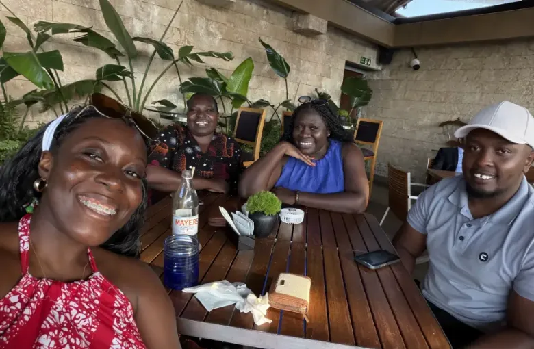 Four colleagues smiling at an outdoor restaurant table in Nairobi; Kenya; with a water bottle and small plant centerpiece on the wooden table.