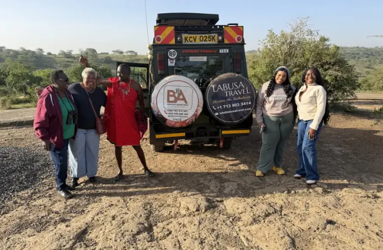 Four travelers pose with a Maasai guide behind a LABUSA Travel safari vehicle on a dirt road in Nairobi Kenya.