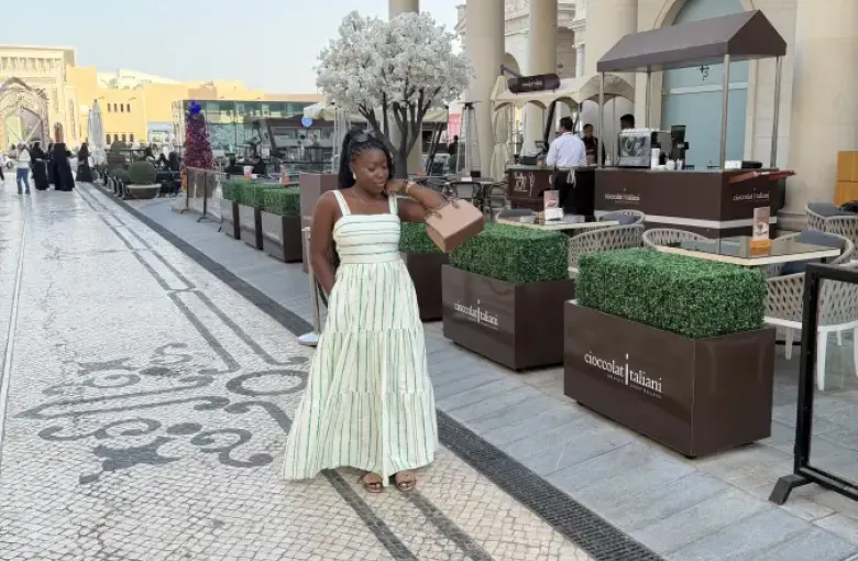 Woman in a striped maxi dress carrying a handbag strolls past Cioccolat Italiani outdoor café in Doha Qatar.