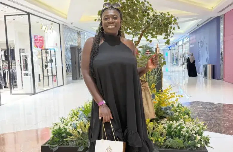 A smiling woman in a black maxi dress holds an Oud Al Sheikh shopping bag and perfume bottle inside a modern mall in Doha Qatar.