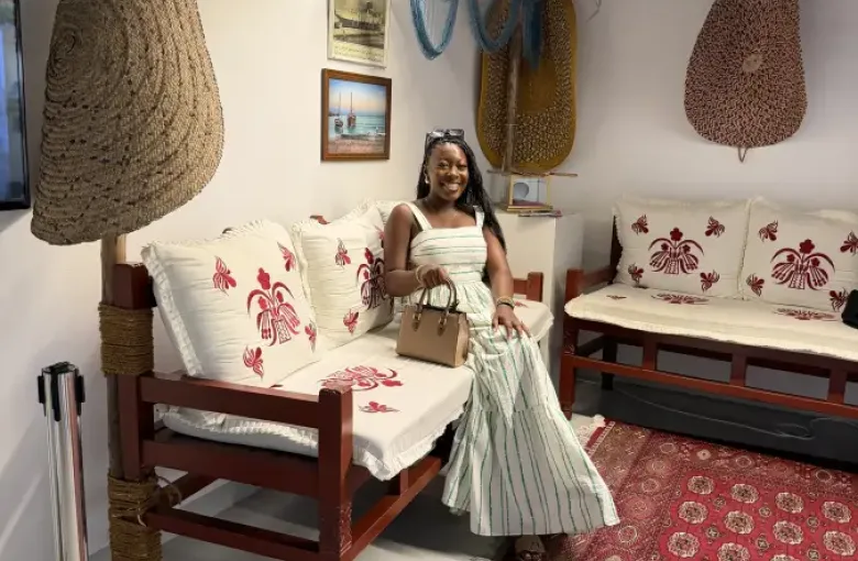 Woman in a striped maxi dress seated on an ornate wooden bench in a traditional Qatari majlis in Doha; decorated with fishing nets and woven baskets.