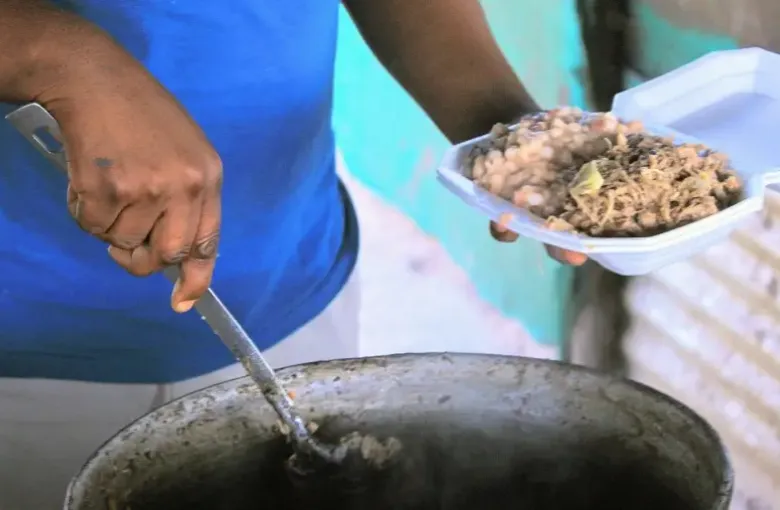 A vendor serves seswaa and dikgobe from a large cooking pot into a takeaway container.