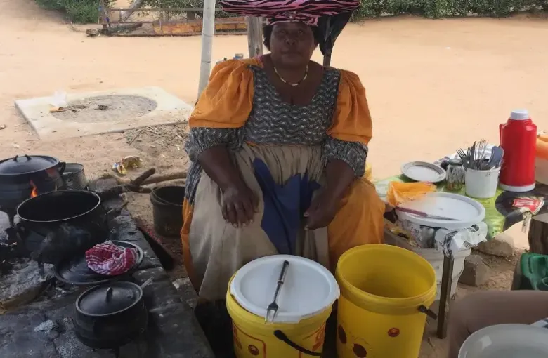 A vendor in Herero dress cooking fat cakes at a roadside stall with pots and buckets.
