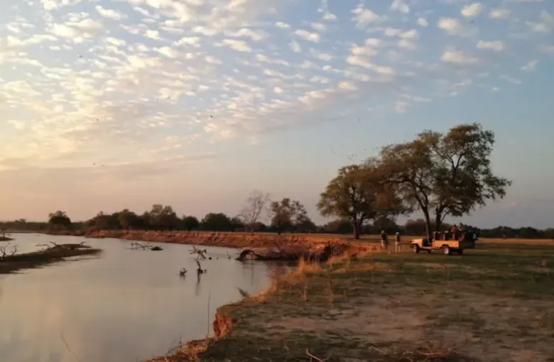 Dawn light over a Zambian river floodplain with scattered trees and a wide cloud-streaked sky.