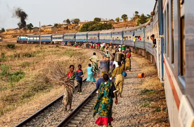 Passengers and traders gather beside a stopped passenger train on a rural railway line.