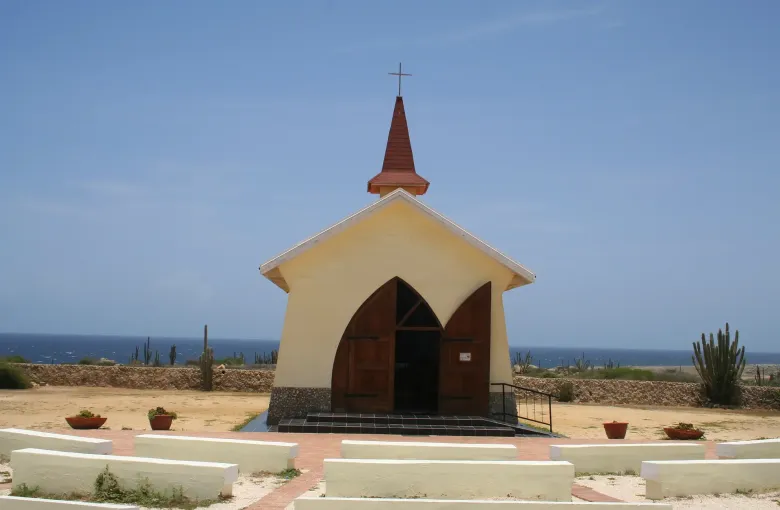 The small yellow Alto Vista Chapel on Aruba, cacti and open sea behind it under a clear sky.