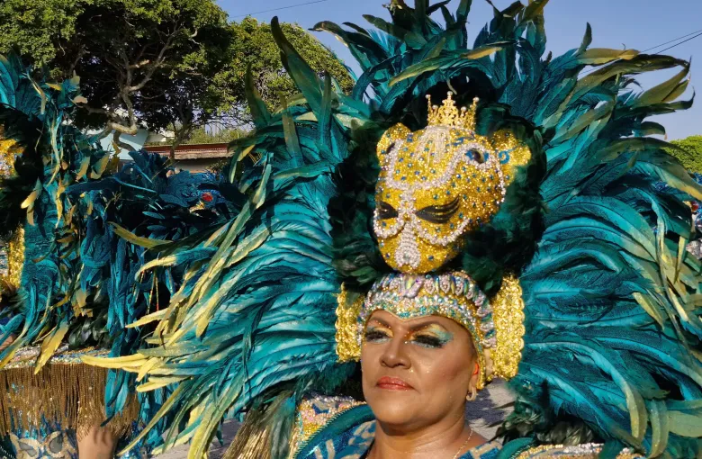 A carnival performer in a teal and gold feathered headdress and jewelled mask at a parade in San Nicolas.