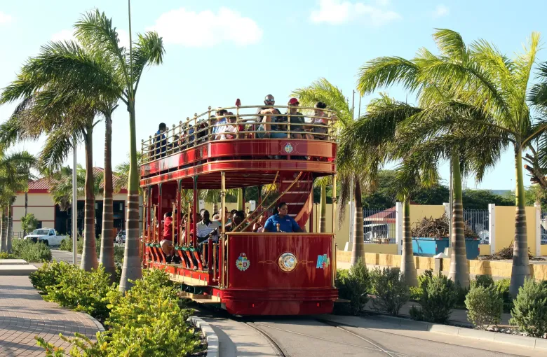 A red open-top double-deck trolley running on rails between palms in central Oranjestad, Aruba.