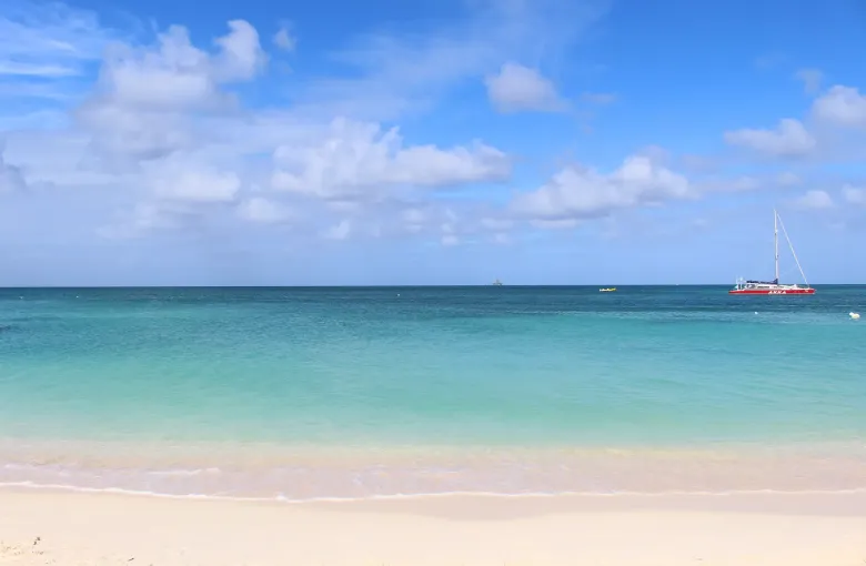 An empty stretch of pale sand at Palm Beach, Aruba, with a catamaran anchored offshore in turquoise water.