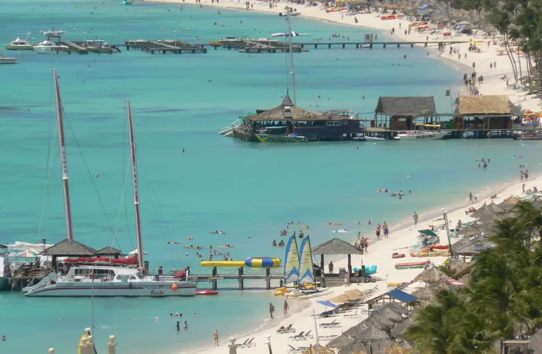 A busy Aruba resort beach from above: catamarans, piers, thatched shade and swimmers in shallow water.