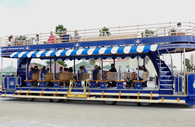 A blue open-sided streetcar with a level floor and a striped canopy on a street in Oranjestad, Aruba.