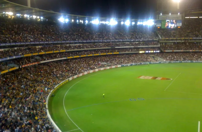 A floodlit Australian Rules football match seen from high in the stands of a packed stadium.