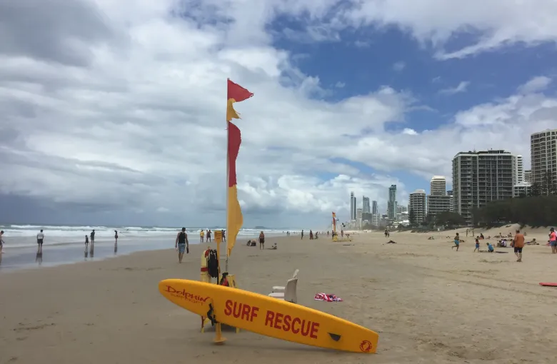 A red and yellow patrol flag and a surf rescue board on the sand, with swimmers beyond them.