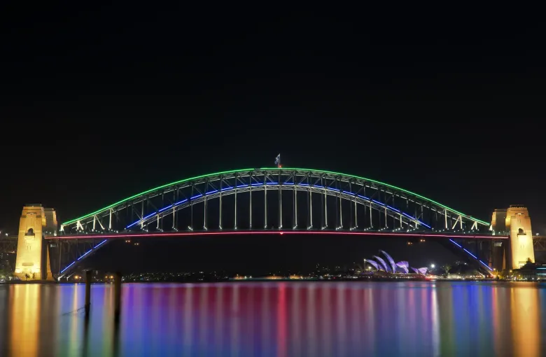 The Sydney Harbour Bridge lit in colored light during the Vivid Sydney festival, reflected in the water.