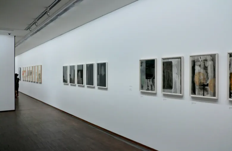 A long white gallery wall in the Albertina in Vienna hung with framed works on paper, one visitor at the far end.