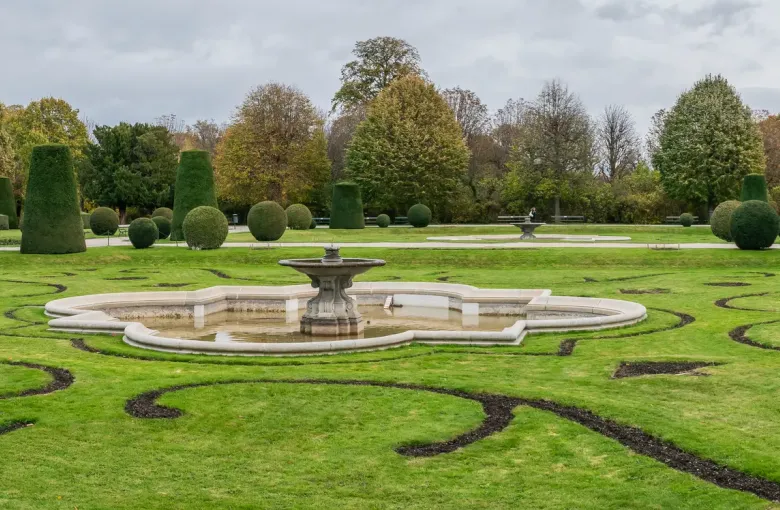 A clipped parterre and fountain in the gardens of Schoenbrunn Palace in Vienna under a gray and white sky.