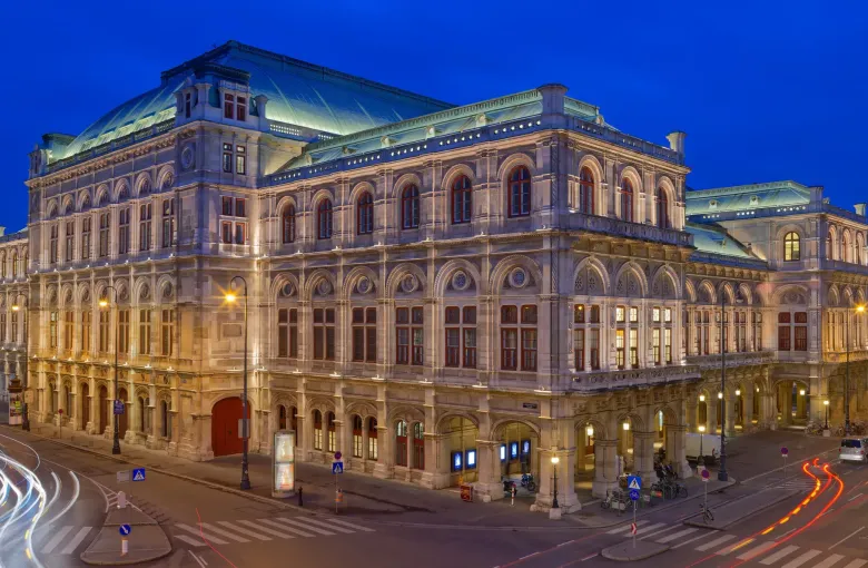 The lit facade of the Vienna State Opera at blue hour, with the wet carriageway of the Ringstrasse in front of it.