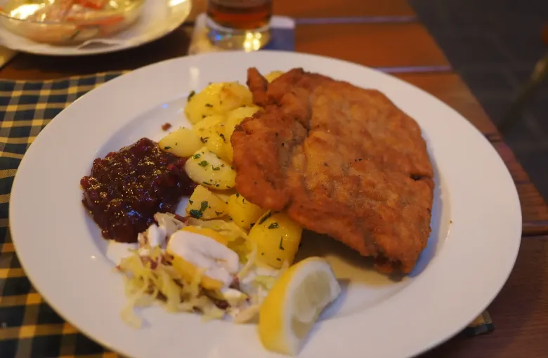 A Wiener Schnitzel served on a white plate with parsley potatoes, lingonberry preserve and a wedge of lemon.