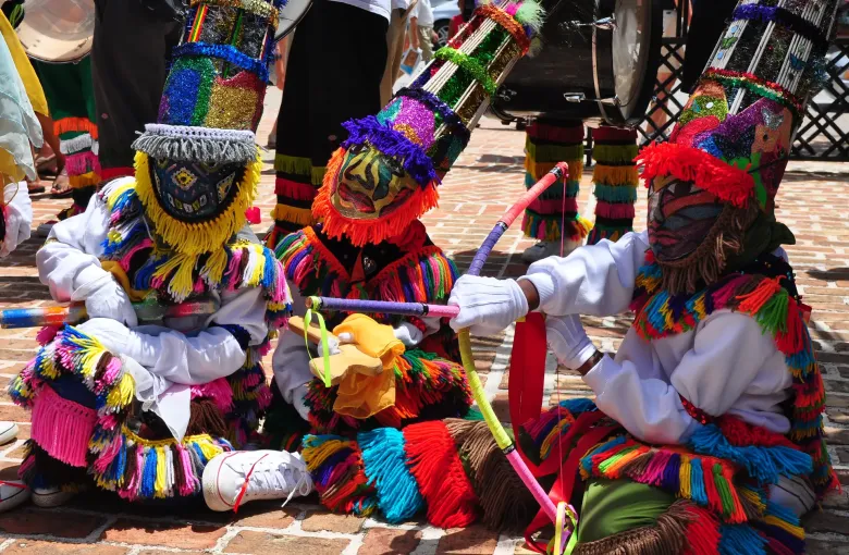 Gombey dancers in fringed capes, painted masks and tall feathered headdresses, a drum behind them.