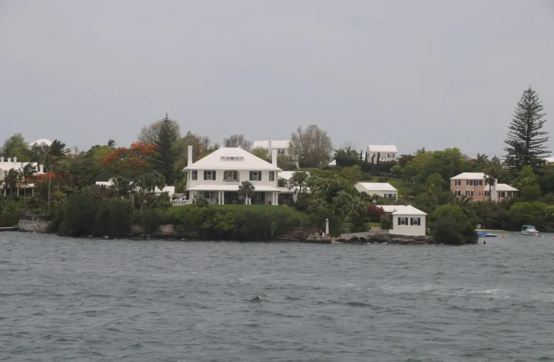 A large white Bermudian house with a stepped roof and dark shutters on a headland above gray water.