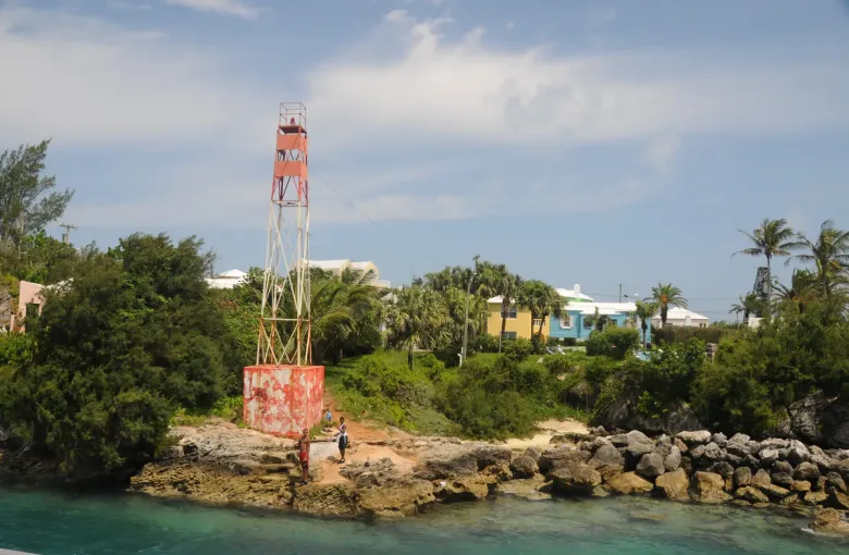 A red navigation beacon on the rocky shore at the Town Cut, pastel houses and palms behind.