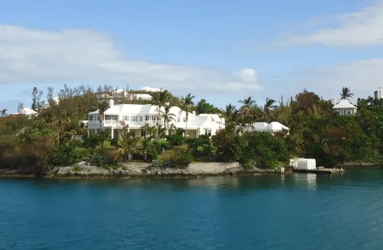 White Bermudian houses with stepped roofs on a wooded headland above clear water.