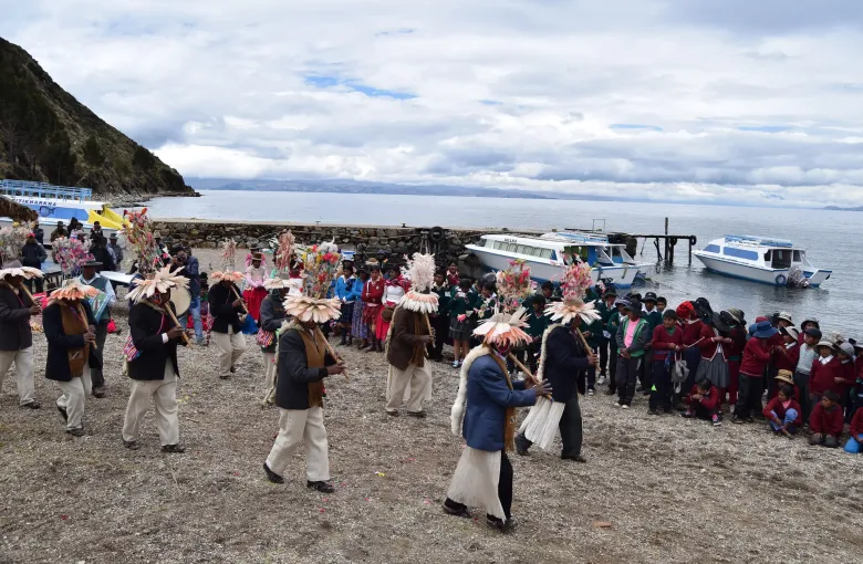 Musicians in feathered headdresses play cane flutes on the shore of Isla del Sol as islanders and schoolchildren watch.