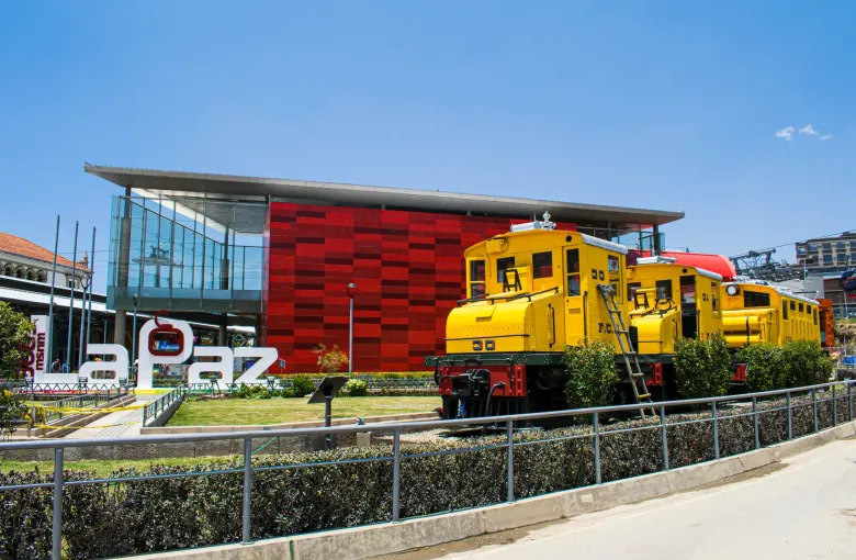 The red clad Estacion Central cable car terminal in La Paz beside preserved railway locomotives and a level paved approach.